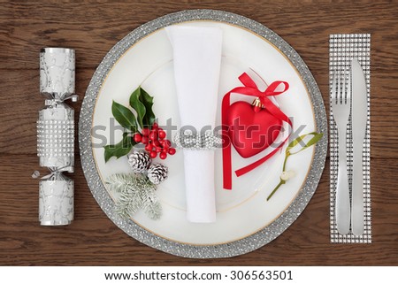 Christmas holiday dinner place setting with plate, napkin, cutlery, sparkling silver and red bauble decorations with holly, mistletoe over oak table background.