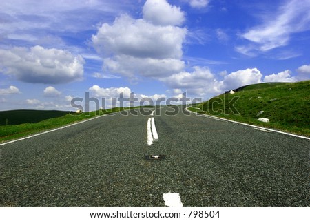 The view of a mountain road  with a right hand bend in the distance against a blue sky with white clouds.
