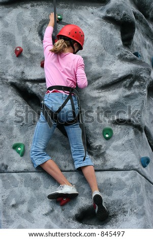 A young girl wearing a safety harness and red hard hat, climbing on a training rock face.