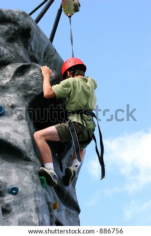 A boy climbing and stretching and nearly at the summit, on a training rock face, wearing a harness and red hard hat.