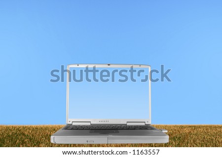 Silver laptop with the lid open and a pale blue screen,  in a field of rough stubble grass against a clear blue sky.