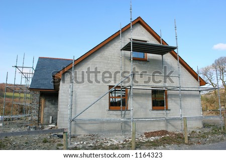 Side view of a house under construction with scaffolding erected to the front and side, set against a blue sky.