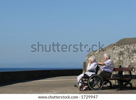Elderly woman in a wheelchair with her hand on her head, seemingly depressed, and sitting next to an elderly man. Sea view and a blue sky in the background.