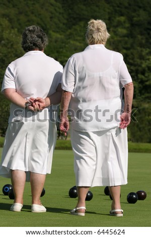 Two elderly ladies dressed in white outfits,  standing together on a lawn bowling green. Rear view.