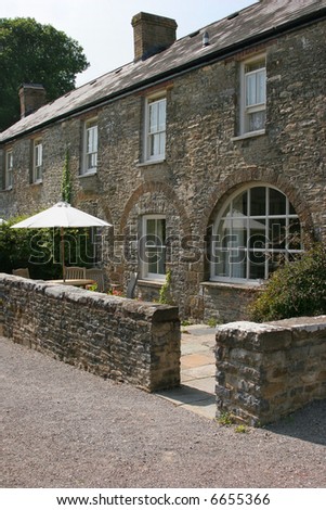 Front of an old stone house with an outdoor patio area, with a wooden table, chairs and a sun umbrella, standing behind a stone wall.