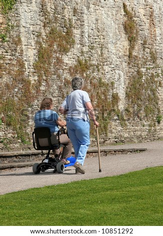 Two disabled females, one sitting on an electric mobility scooter and the other walking alongside with a walking stick. Grass lawn to the foreground and an old stone wall to the rear.