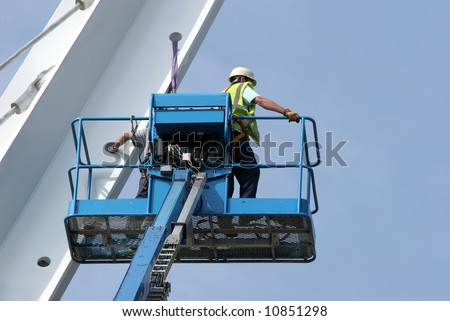 Blue metal cherry picker with workers partially in view,working on a small section of a bridge girder, set against a blue sky.