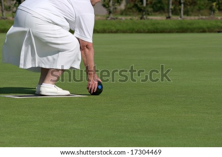 Lower body of an elderly female wearing white clothing and holding a lawn bowling ball about to bowl.