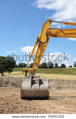 Industrial metal digger hydraulic arm and bucket standing idle on rough earth with rural countryside and a blue sky and clouds to the rear.