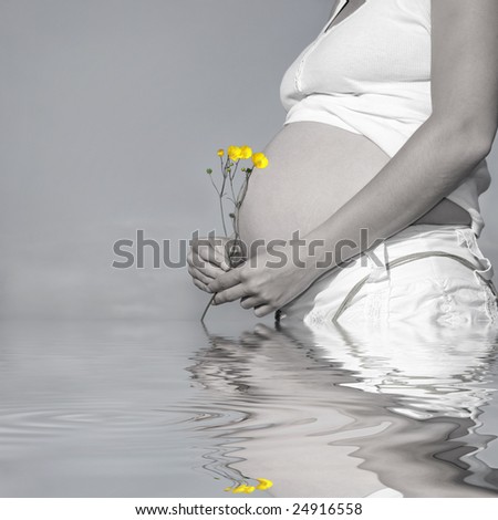 Pregnant woman holding a bunch of buttercups with exposed bare stomach. Desaturated with only the flowers saturated and reflection over rippled water.