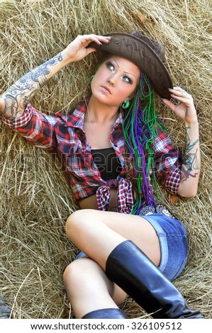 young tattooed  stylish woman with dreadlocks in cowgirl style on haystack background