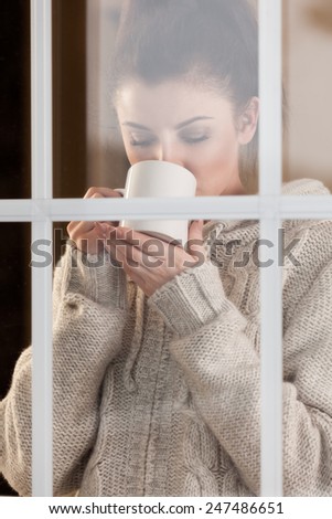 Beautiful woman drinking hot drink standing by the window. View from outside.