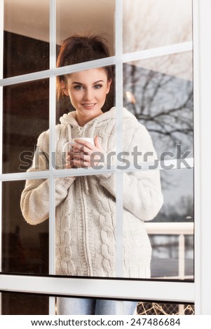 Beautiful woman drinking hot drink standing by the window. View from outside.