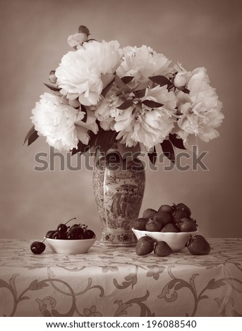 Still Life in style of aged faded photo. Gorgeous white peonies in a porcelain vase with decorative Chinese painting on openwork tablecloth and dishes with sweet strawberries and juicy dark cherries