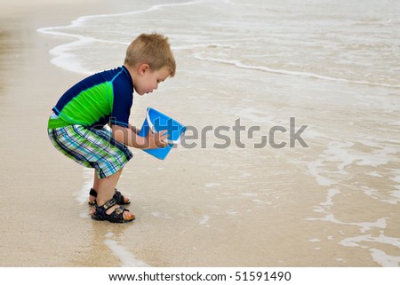 A little boy in a colorful blue and green outfit is playing with a blue bucket at the edge of the water on the beach