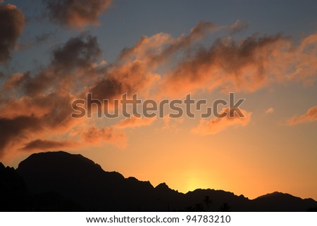 A sunset scene, with brilliant orange yellow and pink clouds against a dark blue sky, with the sihouette of hills where the sun is going down.