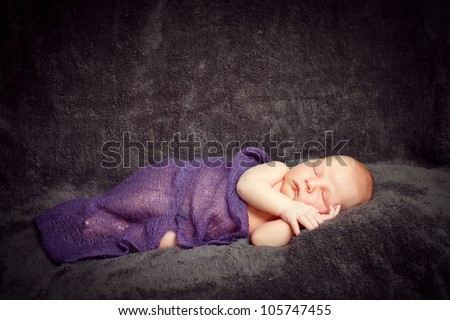 A newborn baby boy is sleeping on a dark background with a blanket. He is wearing a hat. Use it for a childhood, parenting or innocence theme. Soft focus.