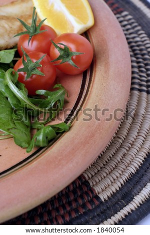 A  glazed stoneware platter or plate of salad on an African woven mat. Some grilled fish in the background.