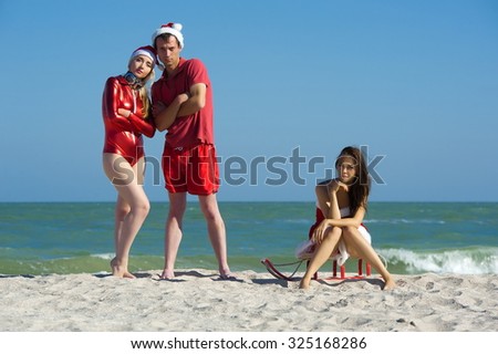 Concept: New Year on the beach. The company - a boy and two girls in clothes for the new year with a sled on the beach