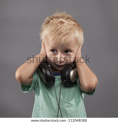 A young boy with headphones around his neck holds his hands over his ears.