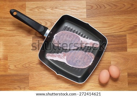 Rashers of raw bacon placed in a griddle frying pan, two brown hens eggs placed at the side on a wooden table top. Shot from above