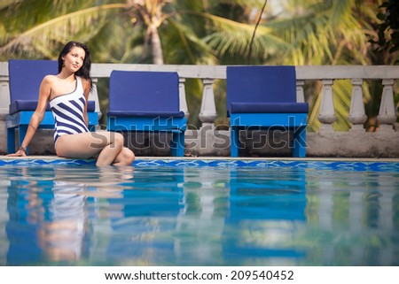 brunette girl in black and white striped swimming suit sitting and posing in blue water in swimming pool with green palms on background