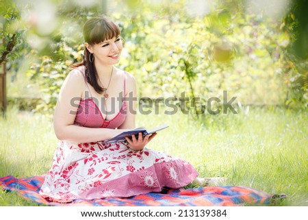 young beautiful brunette girl in pink dress sit on green grass in the garden smile and read a book