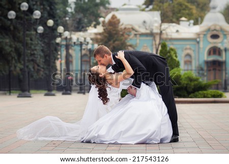 young handsome groom in black suit kiss his beautiful brunette bride in white wedding dress on big city background