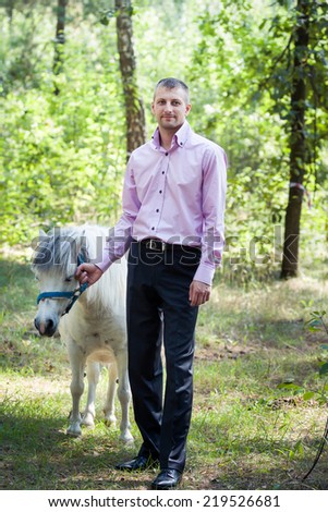 handsome man in pink shirt walk with white pony horse in green forest