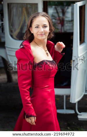 charming young beautiful brunette girl in long red dress look at the camera with white coach on background