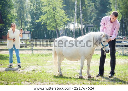 handsome man in pink shirt play with white pony horse on green field with young girl on background