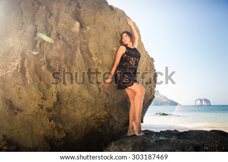 young brunette girl in black transparent lace dress leans pose against large rock