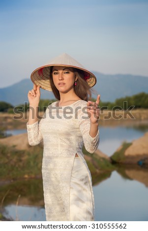 slim blonde girl in Vietnamese national white long dress and hat smiles against lakes country house and mountains