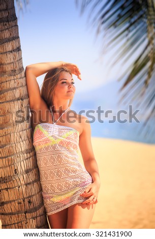 blonde longhaired slim melancholy girl in short white lace frock closeup leans with side elbow on palm trunk on beach