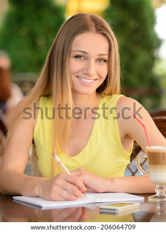 Young beautiful woman drinking coffee and writing in notepad while sitting in a cafe outdoors