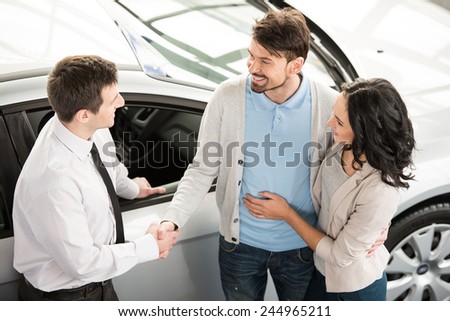 Handsome young car salesman isnstanding at the dealership telling about the features of the car to the couple.