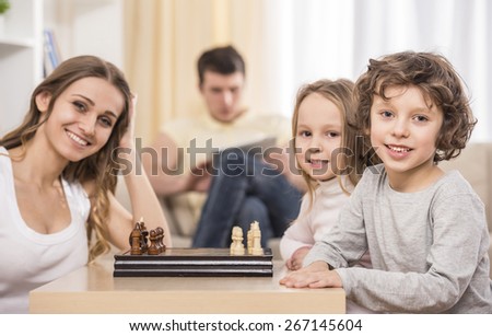 Mother and kids playing chess in living room. Father using a tablet on the couch.
