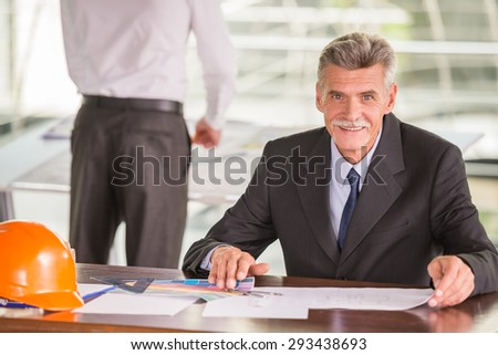 Experienced architect sitting at office desk with orange helmet.