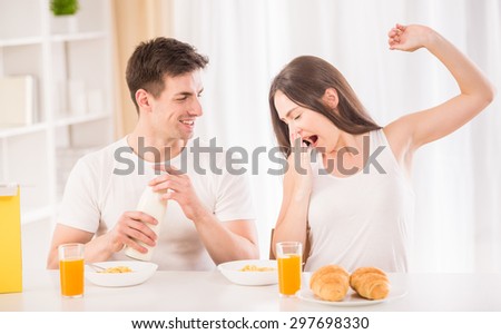 Lazy morning in the kitchen. Young attractive man preparing corn flakes while his sleepy girlfriend making yawn.