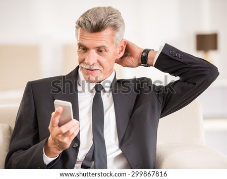 Senior man holding phone and looking on it while sitting at the sofa in hotel room.
