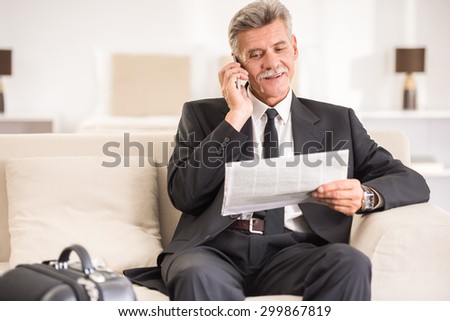 Senior man talking by phone and holding newspaper while sitting at the sofa in hotel room.