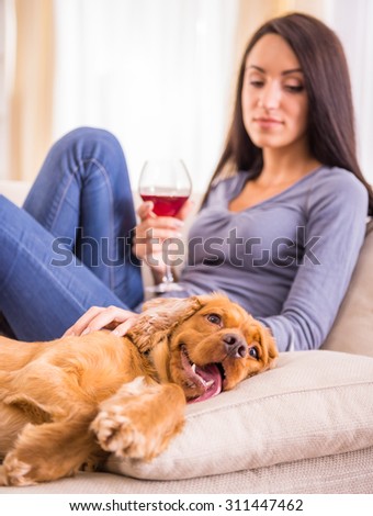 Young woman is relaxing at home with a glass of wine, and her dog is lying near on sofa.