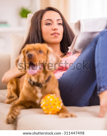 Picture of young woman is reading a newspaper while her dog is lying on sofa near her.