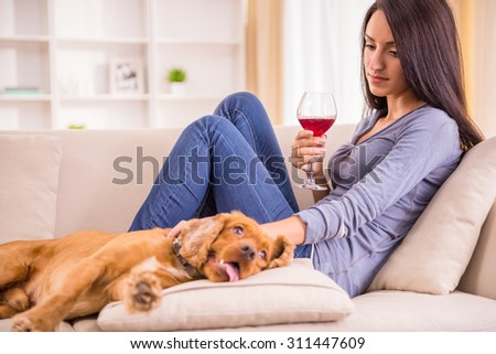 Young woman is relaxing at home with a glass of wine, and her dog is lying near on sofa.