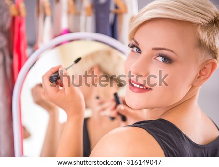 Beautiful young woman is doing makeup in front of the mirror and looking at the camera.