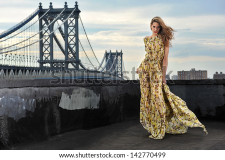 Fashion model posing sexy, wearing long evening dress on rooftop location with metal bridge construction on background