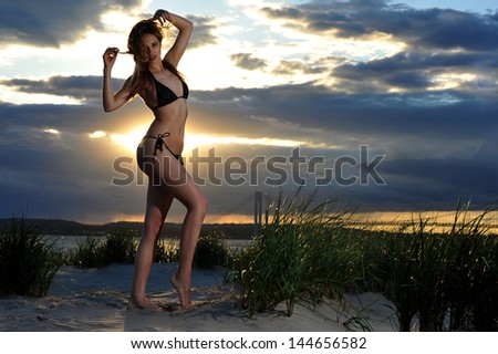 Young fit model posing in bikini on sunset time with effective background of clouds