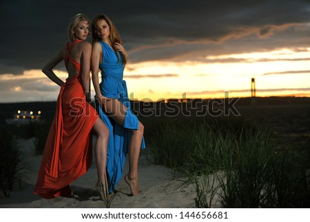 Two fashion model posing in red and blue dresses on sunset time with effective background of clouds and Verrazano bridge
