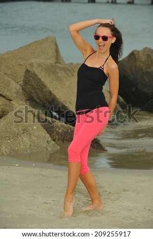 Sporty slim woman in pink leggings and sunglasses posing at the beach with rocks on the background.