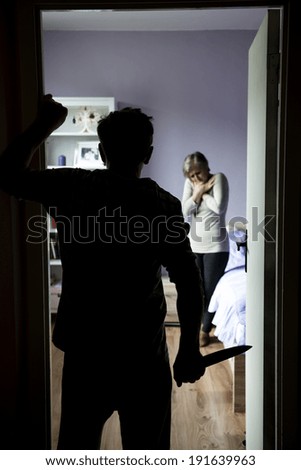 Mature woman in bedroom is scared of man with knife. Woman is victim of domestic violence and abuse.